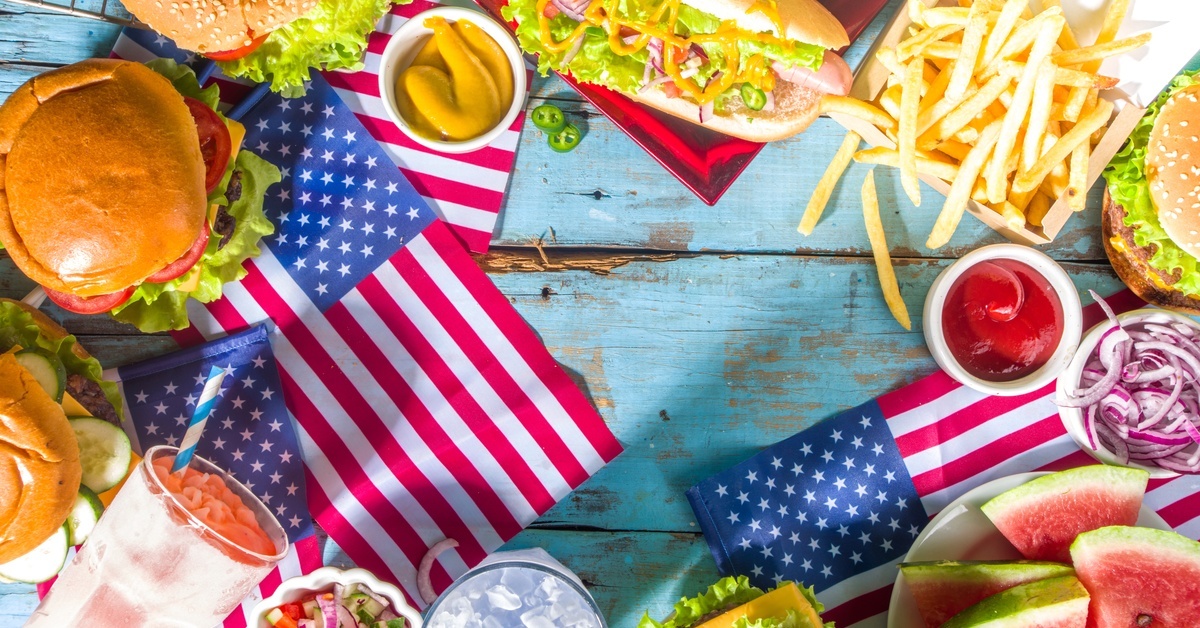 A blue picnic table almost completely covered with food and drinks. A small, North American fly lays in the middle of it all.