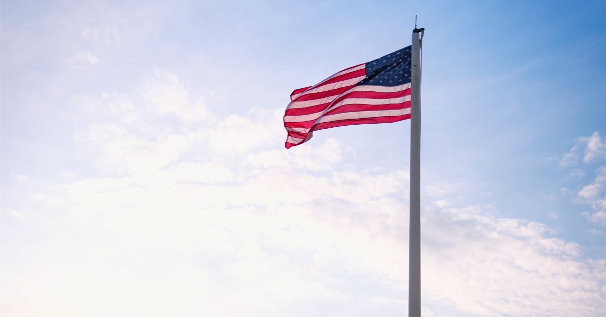 A tall flagpole has a large American flag flying on the top. Past the flag and flagpole there is a sunrise.