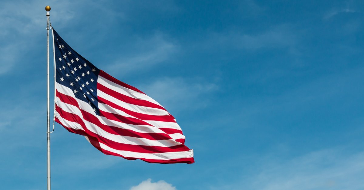 A close up of an American flag hanging on top of a flagpole, waving in the wind. there are clouds behind it.