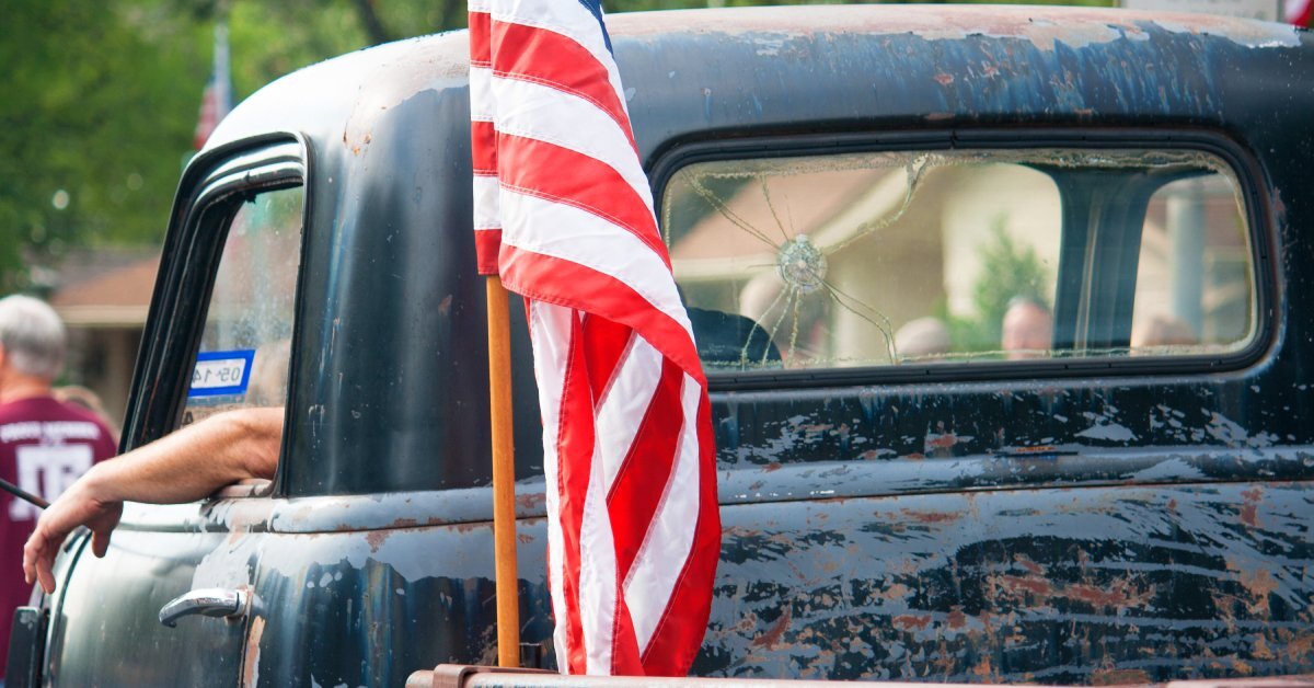 An old beat-up pickup truck has an American flag in the back with only the red and white stripes visible.