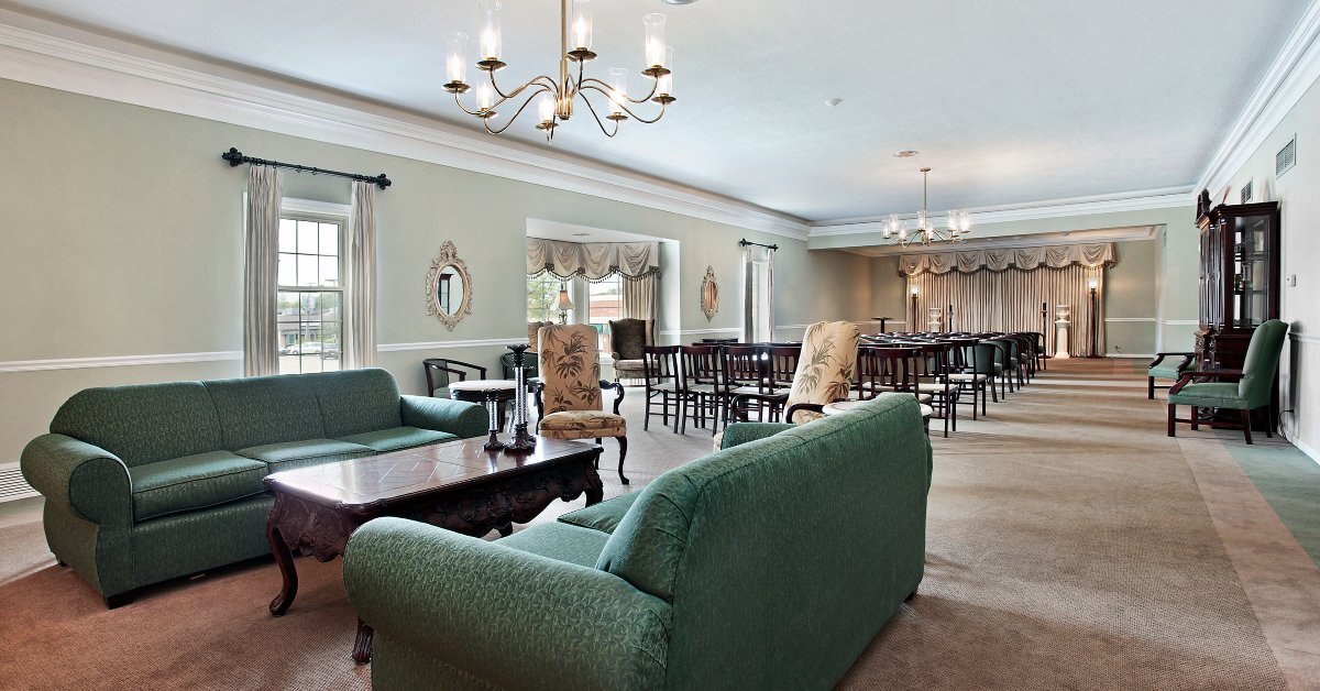A funeral home with numerous chairs, couches, and tables. The room is incredibly long with multiple rows of chairs facing the front of the room.