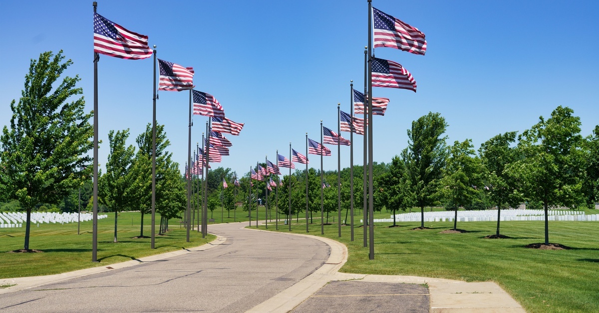 Why US State Flags Make Great Gifts for History Buffs A large cement pathway that has large flag poles with American flags flying high. There's green grass with trees next to the flagpoles.