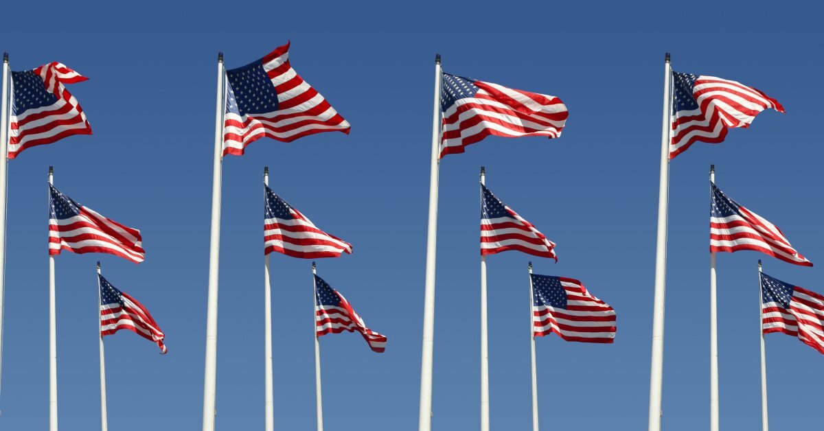 Multiple flag poles standing next to each other against the sky. Each flag pole has an American flag flying.