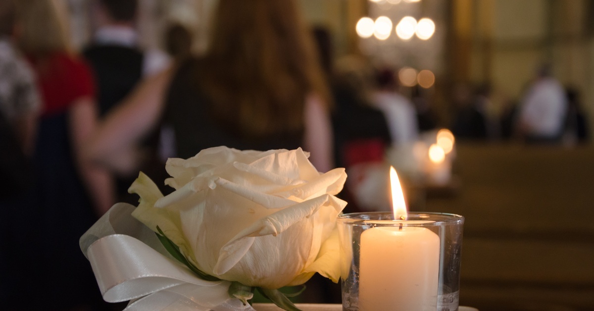 A church service with people in the distance standing in their pews, while the foreground has a flower and lit candle.