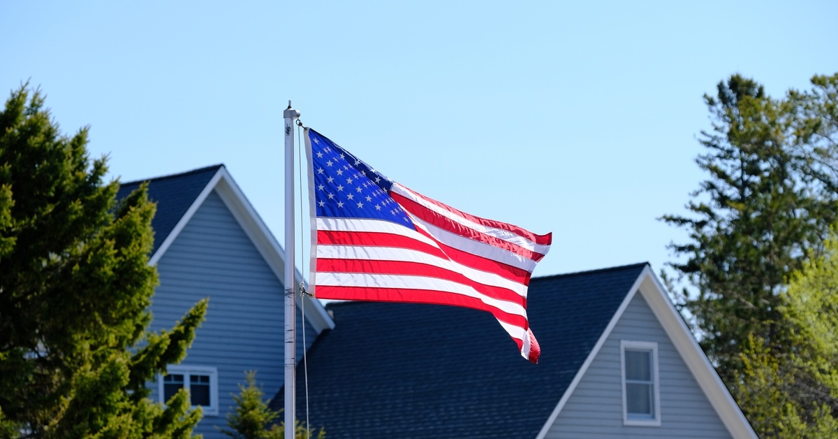 How To Select the Right Height for an In-Ground Flagpole A large blue house sits in between some trees while a bright American flag flies high in front of the home.