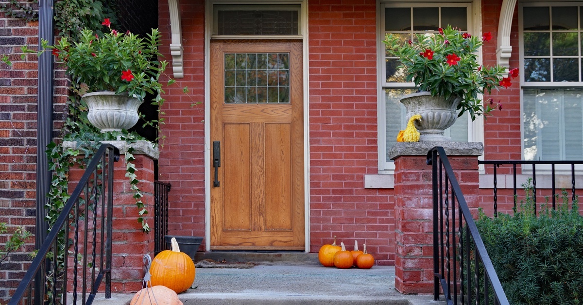 The front porch and door of a red-brick house featuring wrought-iron railings, pumpkins, and potted plants.