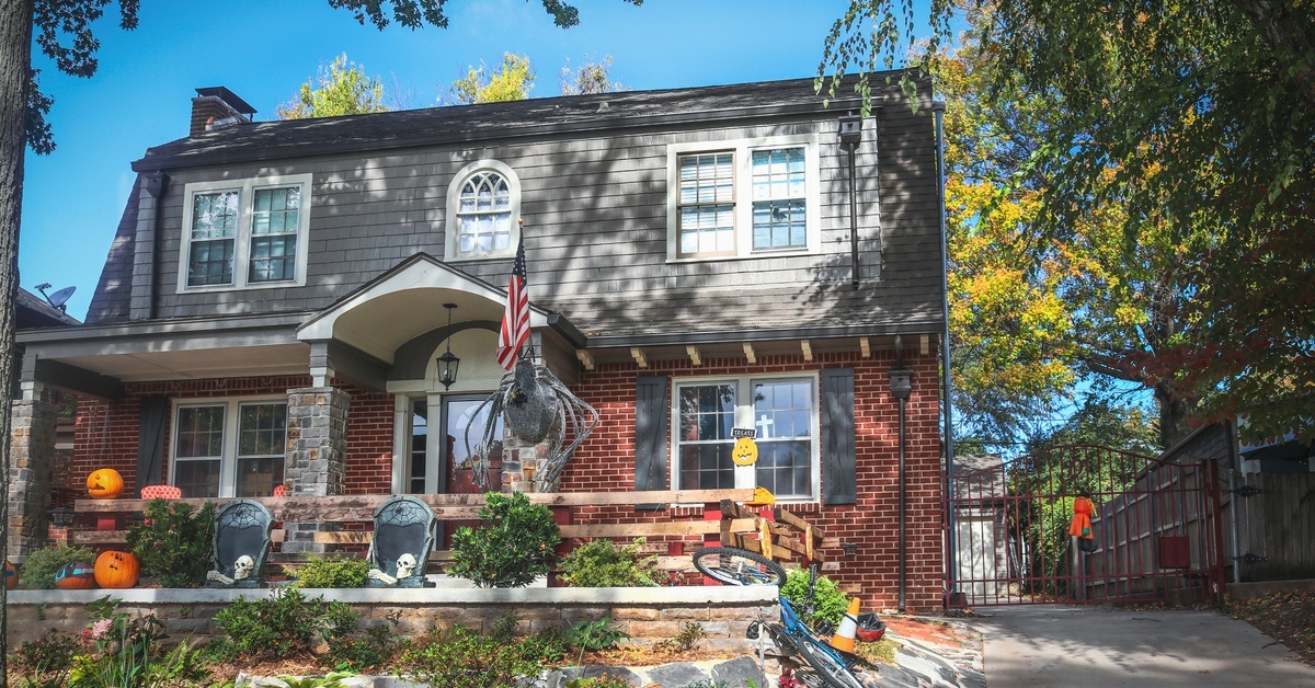 A vintage house with a red-brick facade, front lawn, and large oak tree. Its front porch features Halloween decorations.